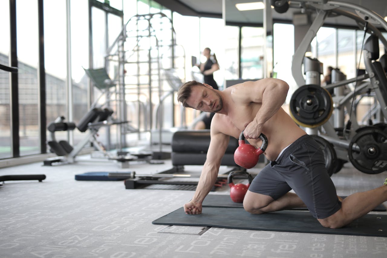 Shirtless man performing kettlebell exercise in a bright, modern gym space.